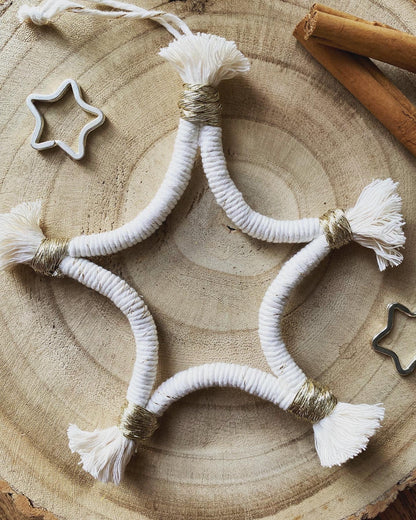 A macrame star shaped hanging decoration, made with natural yarn with gold detailing, and fringed ends, laying on a wooden board. There are cinnamon sticks and metal stars laid out nearby for decoration. 