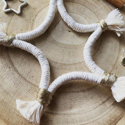 Close up of a macrame star shaped hanging decoration, made with natural yarn with gold detailing, and fringed ends, laying on a wooden board. 
