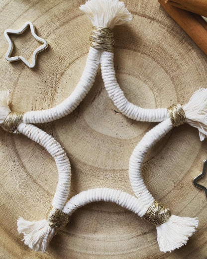 A macrame star shaped hanging decoration, made with natural yarn with gold detailing, and fringed ends, laying on a wooden board. 