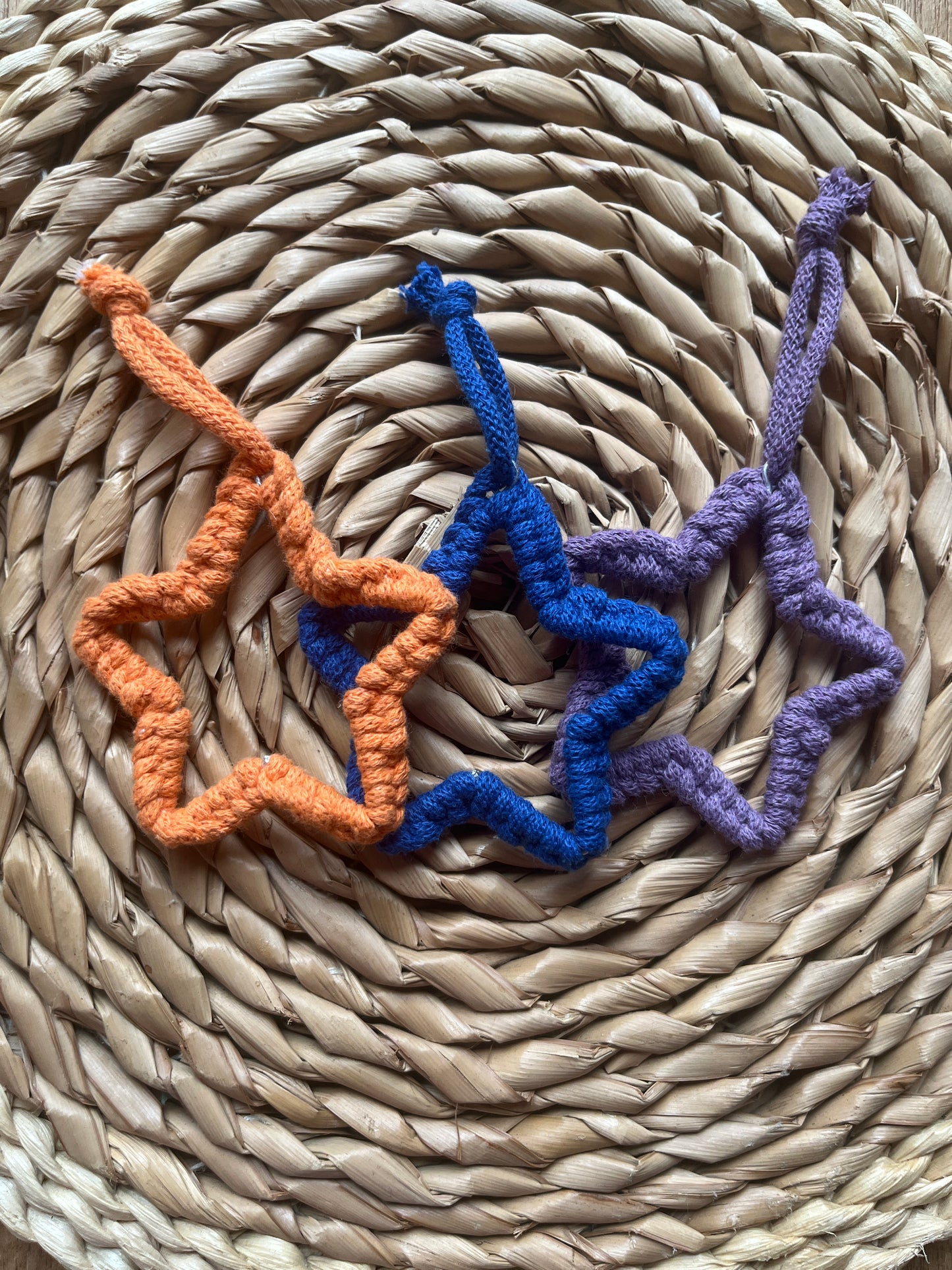 Three macrame star-shaped decorations in purple, blue, and orange on a woven mat.