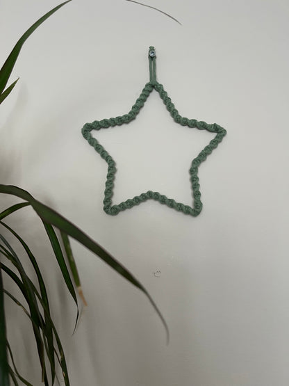 Star shaped wall hanging, wrapped in green cotton with a twisted knot pattern, hanging on a beige wall. There is a green houseplant in the bottom left of the image. 