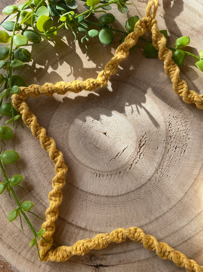 Star shaped wall hanging, wrapped in mustard yellow cotton with a twisted knot pattern, laid flat on a wooden surface with green plant vines. The sun is shining on to the image casting shadows.  