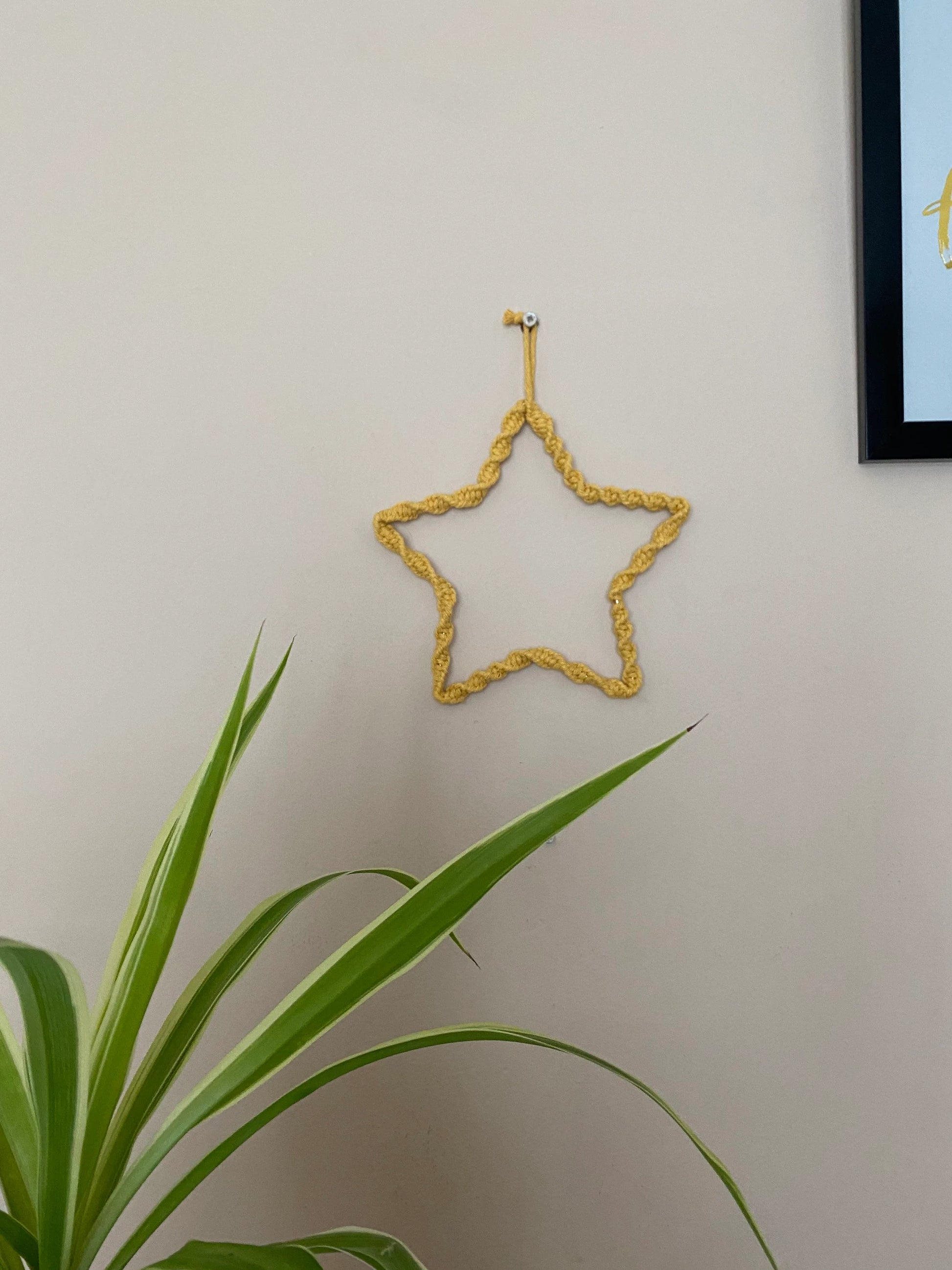Star shaped wall hanging, wrapped in mustard yellow cotton with a twisted knot pattern, hanging on a beige wall. There is a green houseplant in the bottom left of the image. 