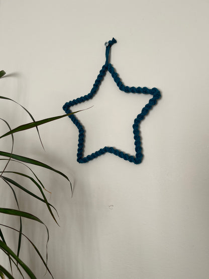 Star shaped wall hanging, wrapped in petrol blue cotton with a twisted knot pattern, hanging on a beige wall. There is a green houseplant in the bottom left of the image. 