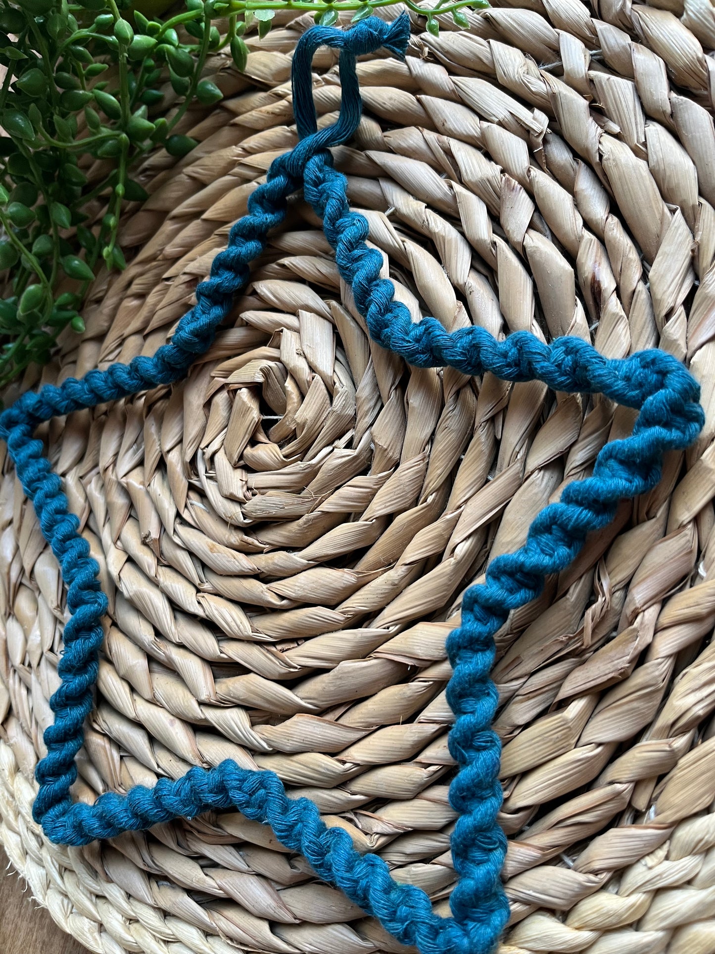 Star shaped wall hanging, wrapped in petrol blue cotton with a twisted knot pattern, laid flat on a woven mat next to some green plant vines. 
