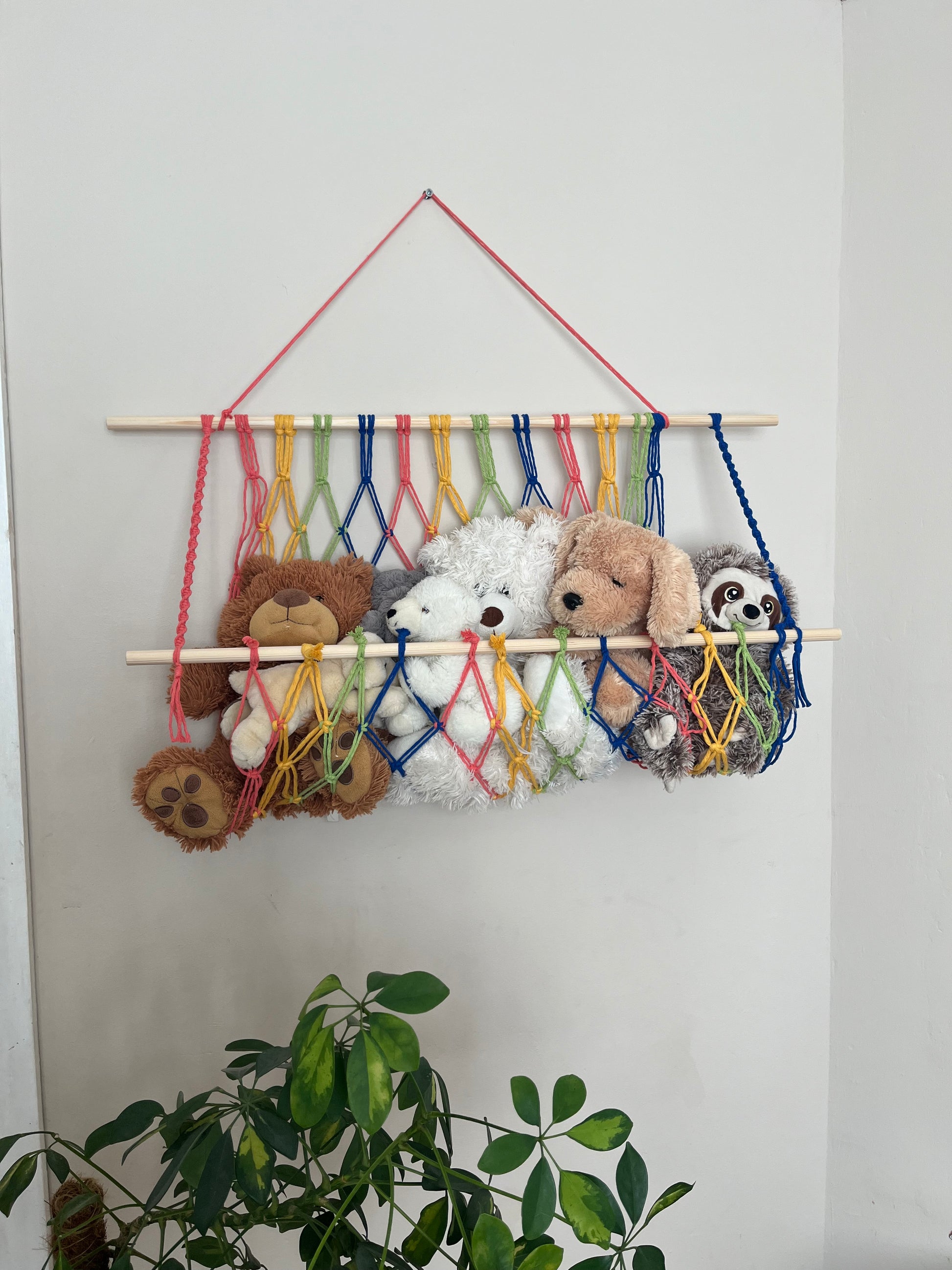 A macrame toy hammock made with colourful yarn and wooden dowels, hanging against a beige wall, filled with soft toys and teddies. There is a large green houseplant just below. 