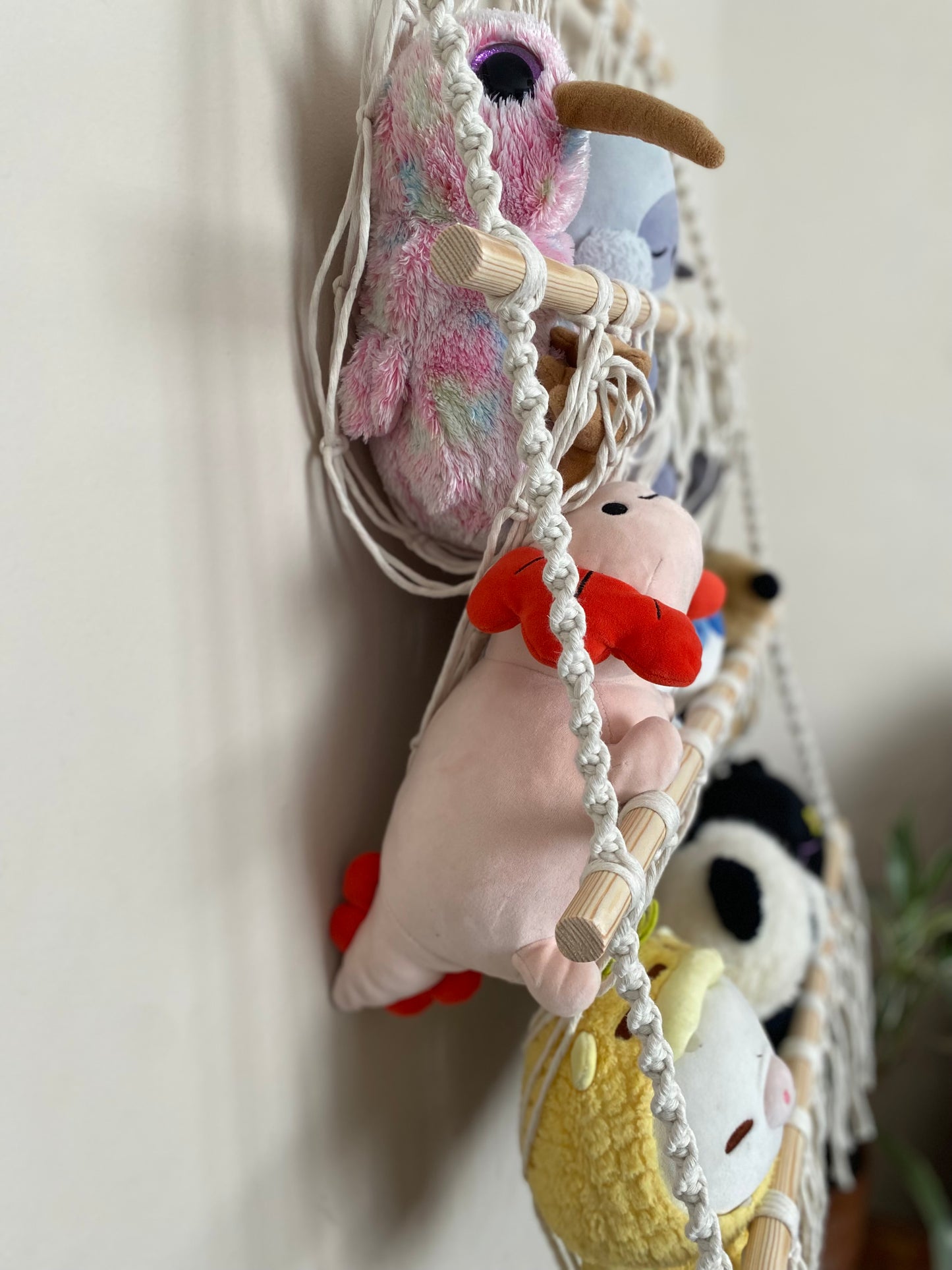 Close up of a macrame toy hammock made with natural yarn and wooden dowels, hanging against a beige wall, filled with soft toys and teddies. 