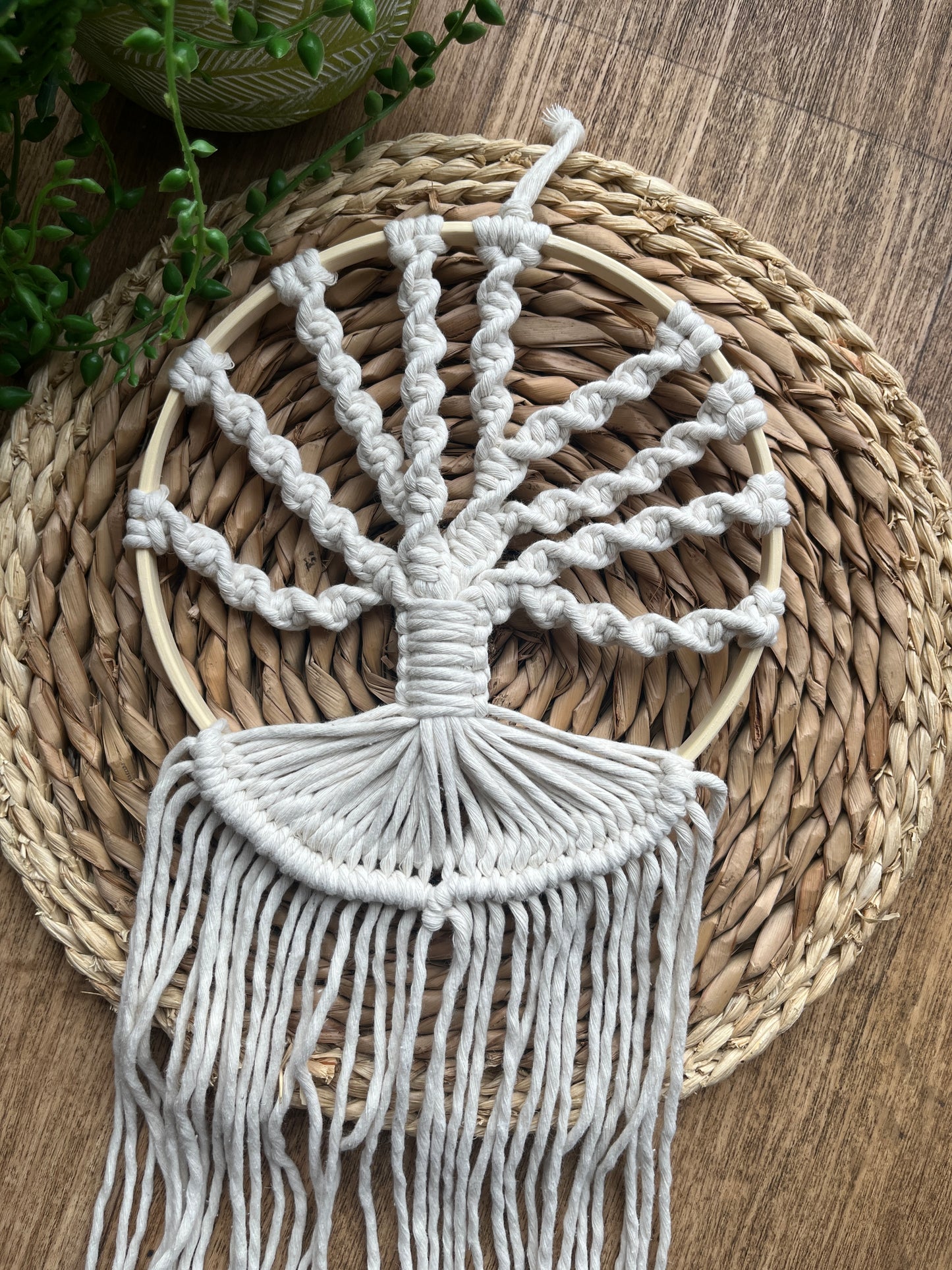 A macrame tree of life wall hanging made on a circular wooden hoop with natural knotted yarn, in the shape of tree branches, placed on a circular woven mat. 