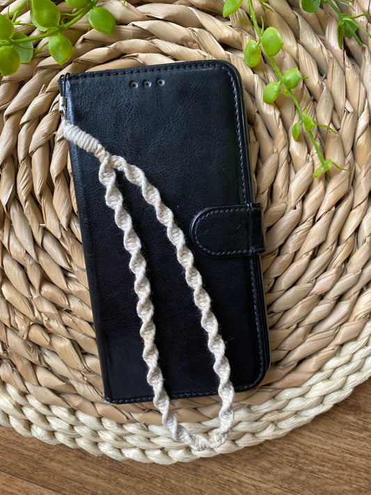 A black wallet style phone case lays on a woven wicker placemat. The phone case has a macrame phone charm made with beige coloured yarn attached to it at the top and the charm is laid across the top of the phone case. 