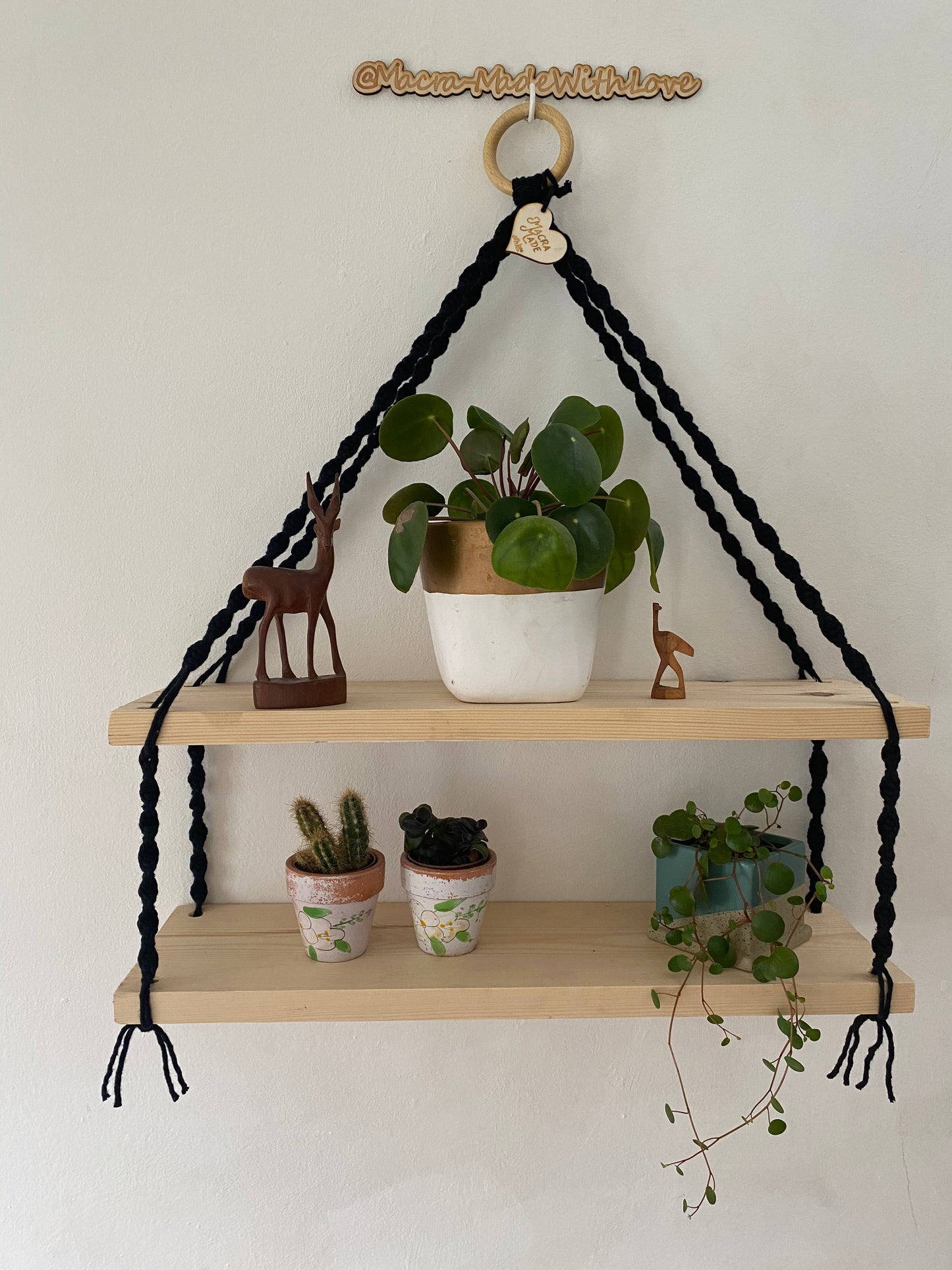 A set of macrame shelves, made with two solid wood shelf pieces, suspended by black knotted yarn, on a wooden hanging ring, hanging against a white painted wall. There are various small plant and wooden ornaments on the shelves. 