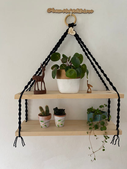 A set of macrame shelves, made with two solid wood shelf pieces, suspended by black knotted yarn, on a wooden hanging ring, hanging against a white painted wall. There are various small plant and wooden ornaments on the shelves. 