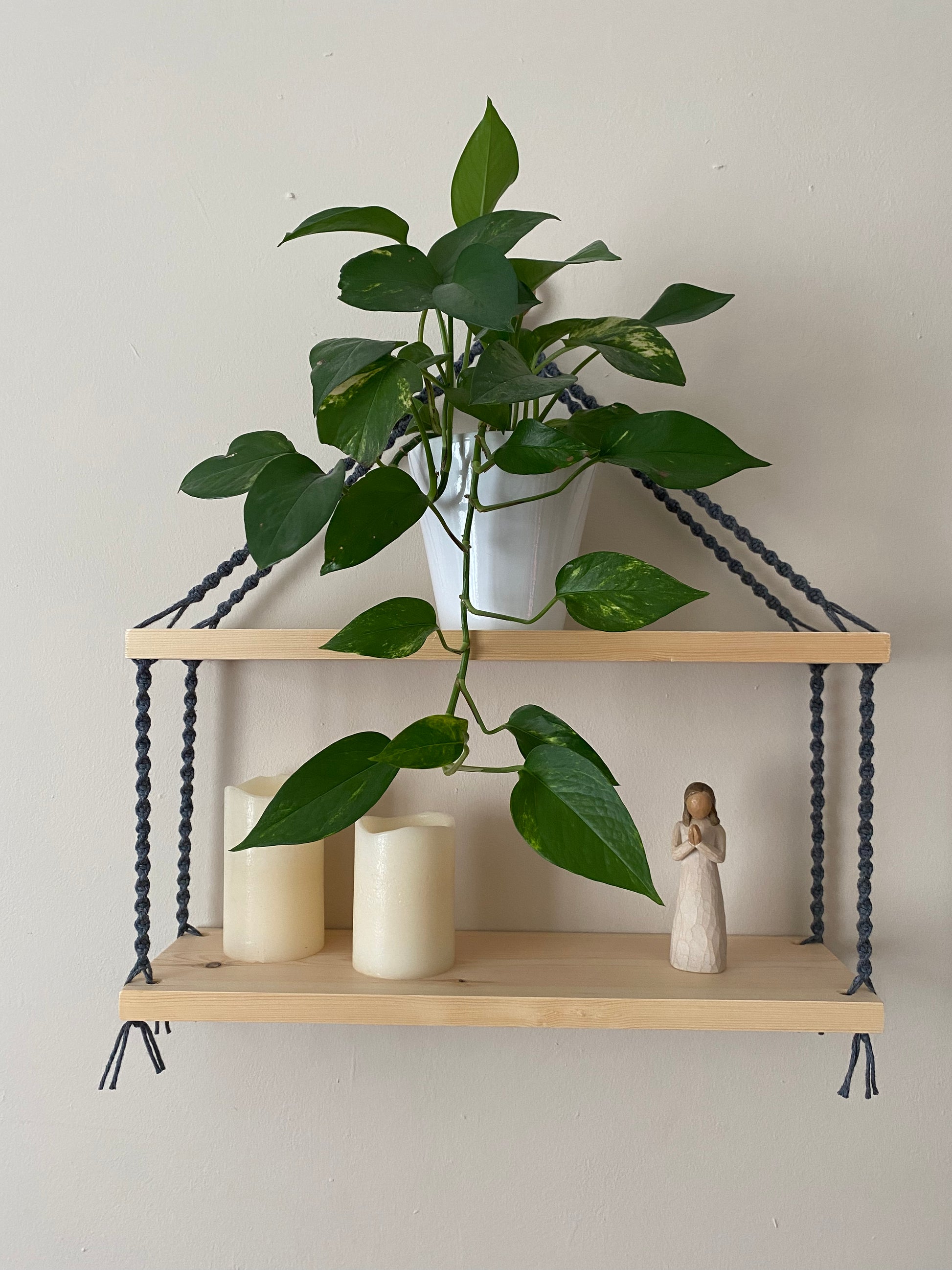 A set of macrame shelves, made with two solid wood shelf pieces, suspended by dark grey coloured knotted yarn, on a wooden hanging ring, hanging against a beige painted wall. There is a long green trailing plant on the top shelf, an ornament and two pilar candles on the bottom shelf.
