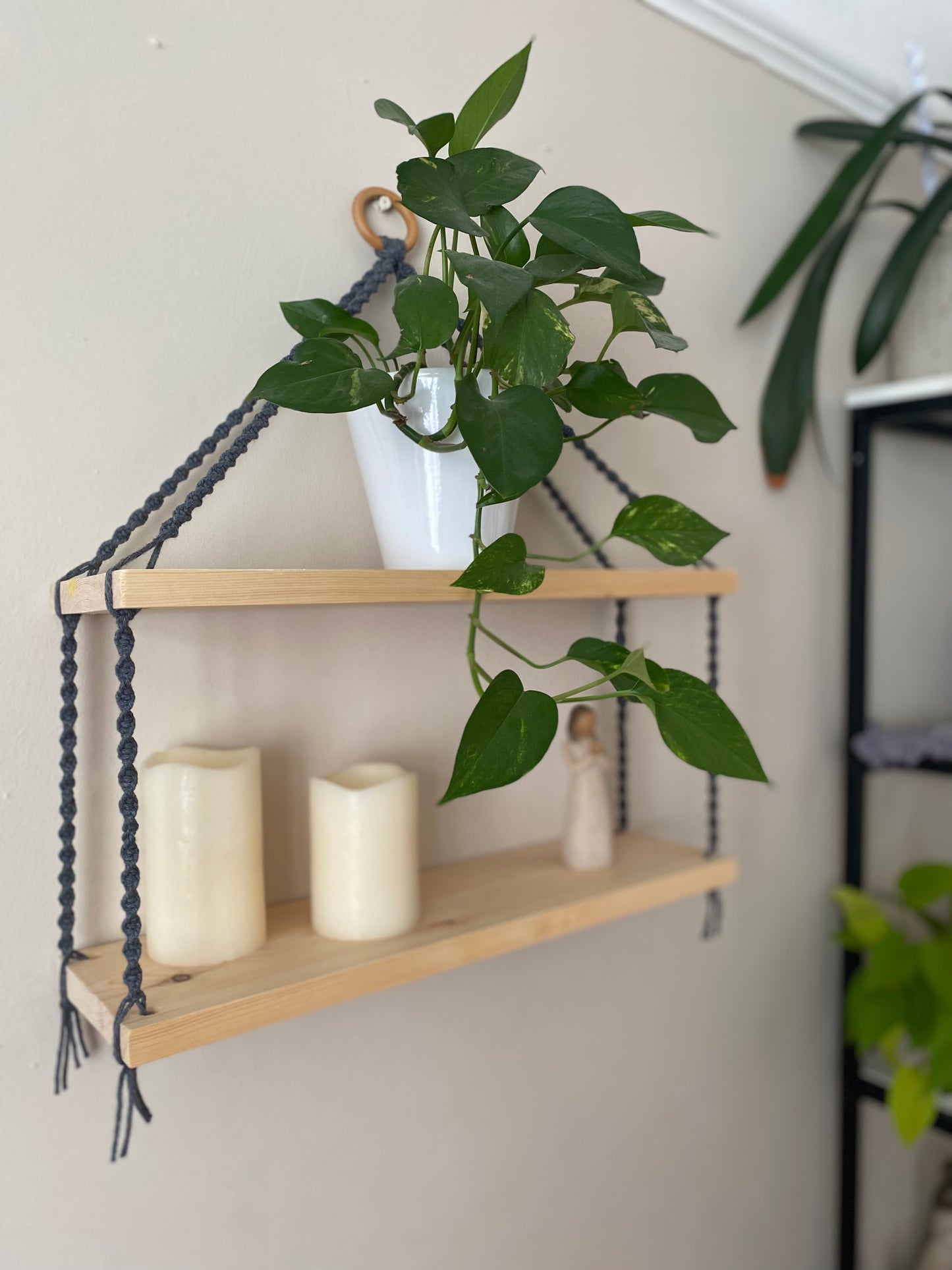 Side angled view of a set of macrame shelves, made with two solid wood shelf pieces, suspended by dark grey coloured knotted yarn, on a wooden hanging ring, hanging against a beige painted wall. There is a long green trailing plant on the top shelf, a small ornament and two pillar candles on the bottom shelf. 