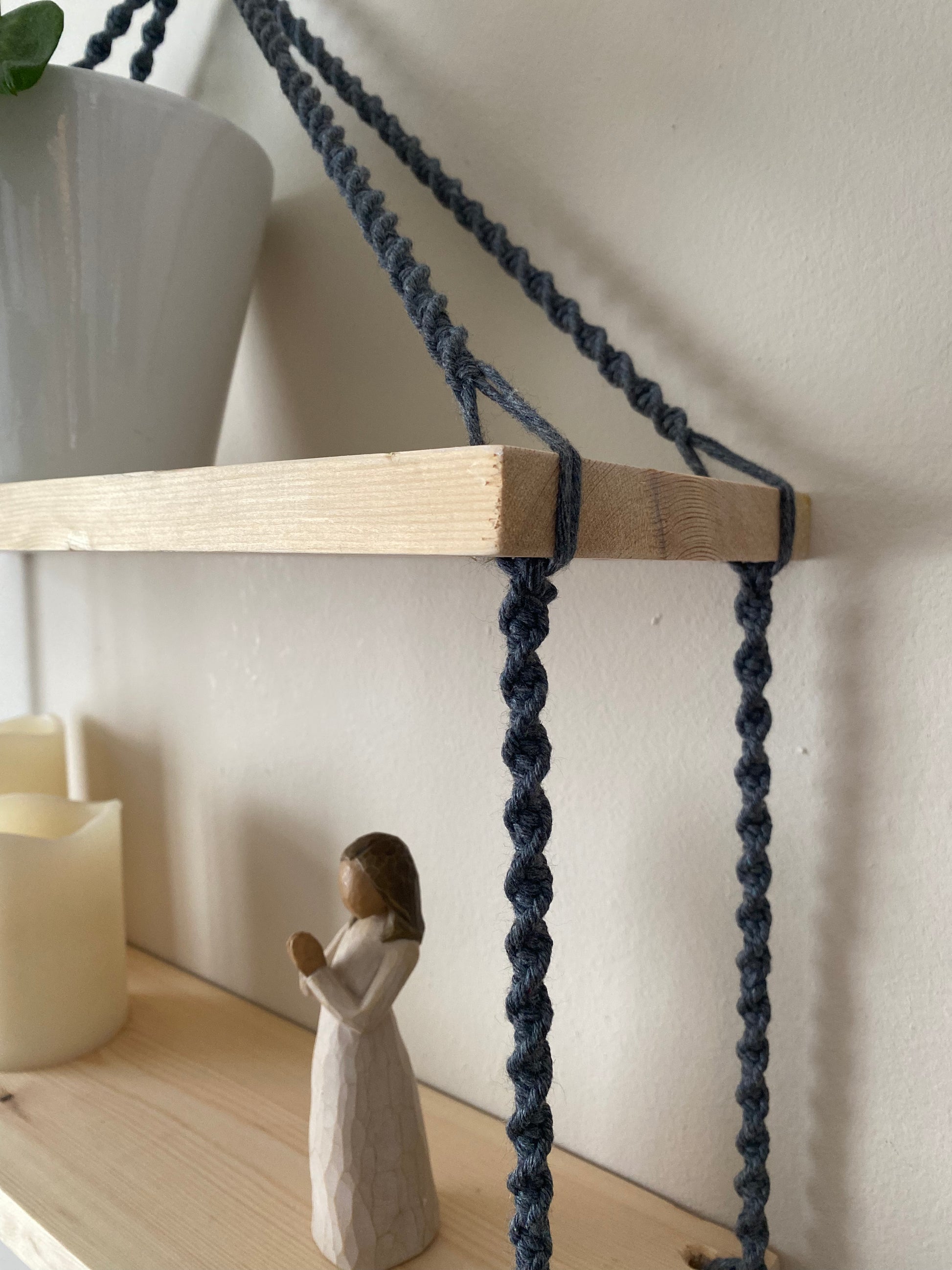 A close up of a set of macrame shelves, made with two solid wood shelf pieces, suspended by dark grey coloured knotted yarn. A plant pot is just seen on the top shelf, and there is a small ornament and pillar candles on the bottom shelf. 