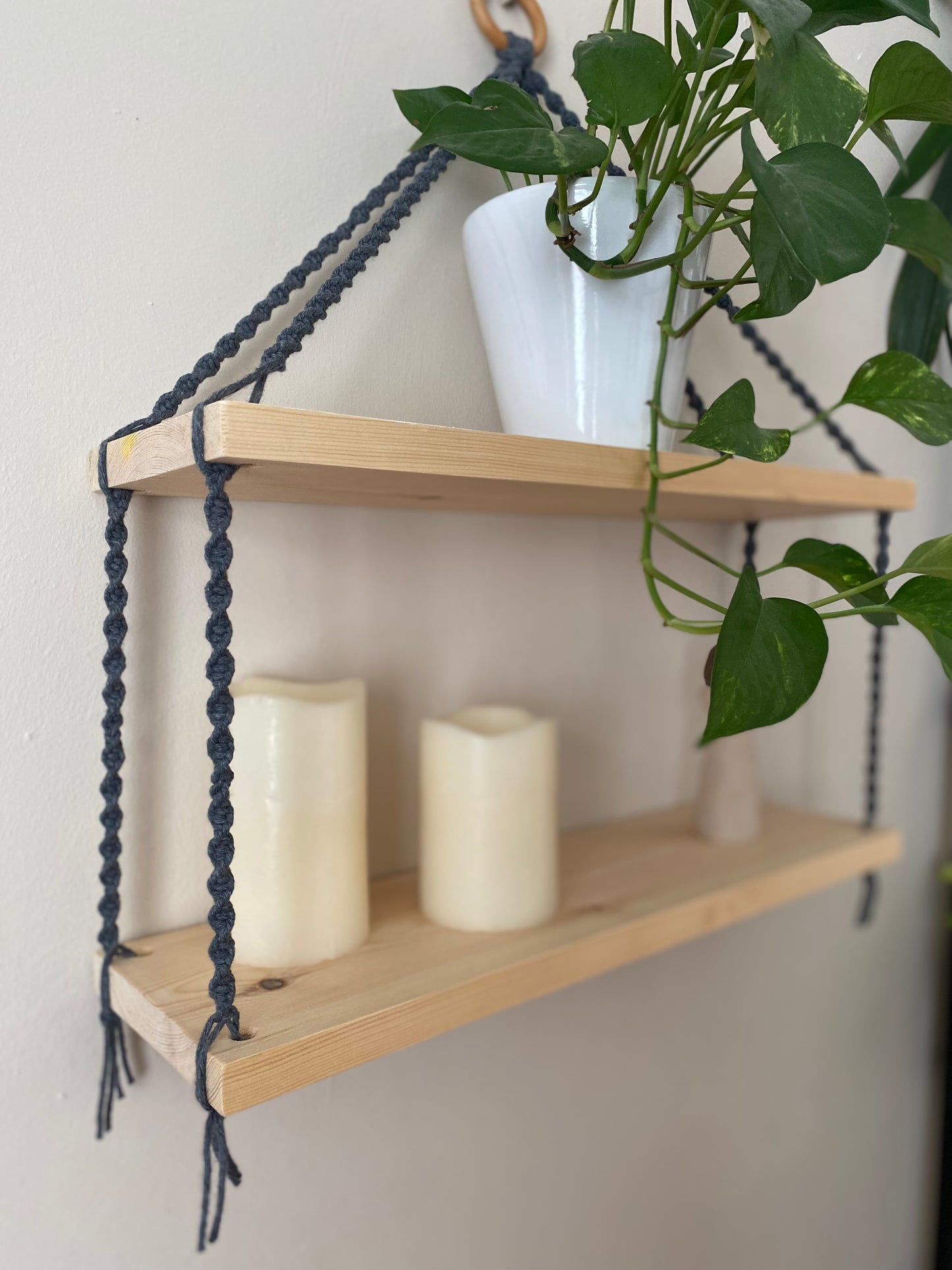 A close up of a set of macrame shelves, made with two solid wood shelf pieces, suspended by dark grey coloured knotted yarn, hanging against a beige painted wall. There is a long green trailing plant on the top shelf, and two pillar candles on the bottom shelf. 