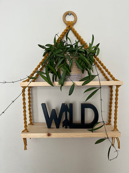 A set of macrame shelves, made with two solid wood shelf pieces, suspended by mustard yellow coloured knotted yarn, on a wooden hanging ring, hanging against a beige painted wall. There is a long green trailing plant on the top shelf, and a wooden sign in black which says "WILD" on the bottom shelf.