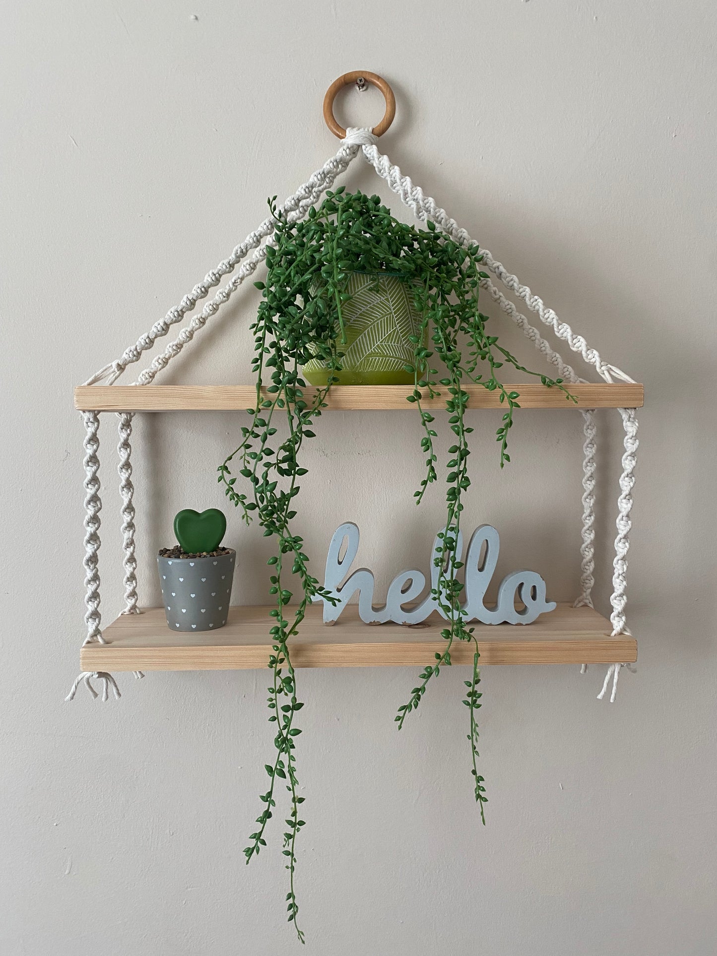 A set of macrame shelves, made with two solid wood shelf pieces, suspended by natural coloured knotted yarn, on a wooden hanging ring, hanging against a beige painted wall. There is a long green trailing plant on the top shelf, a small plant and a wooden sign that says "hello" on the bottom shelf. 