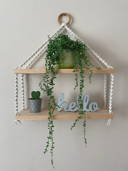 A set of macrame shelves, made with two solid wood shelf pieces, suspended by natural coloured knotted yarn, on a wooden hanging ring, hanging against a beige painted wall. There is a long green trailing plant on the top shelf, a small plant and a wooden sign that says "hello" on the bottom shelf. 