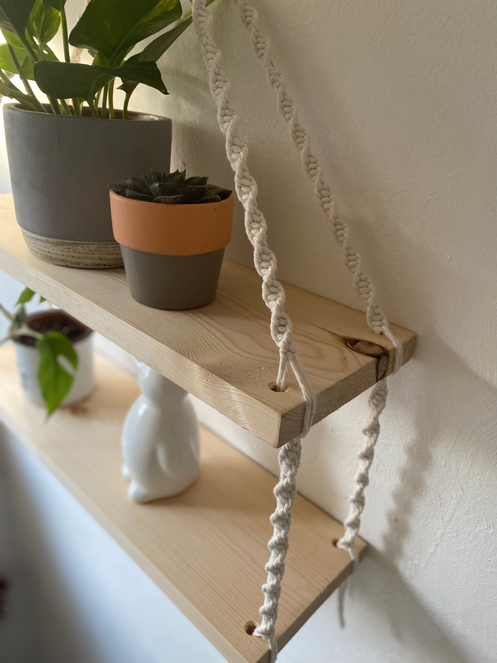 Close up of a set of macrame shelves, made with two solid wood shelf pieces, suspended by natural coloured knotted yarn, hanging against a white painted wall. There are various plants and ornaments on the shelves. 