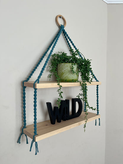 Side angled view of a set of macrame shelves, made with two solid wood shelf pieces, suspended by teal blue coloured knotted yarn, on a wooden hanging ring, hanging against a beige painted wall. There is a long green trailing plant on the top shelf, and a wooden sign in black which says "WILD" on the bottom shelf.