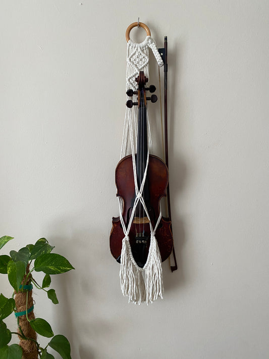 A dark wooden violin and bow hanging in a macrame wall hanger, made with natural off white coloured recycled cotton on a wooden ring, with long boho fringe.