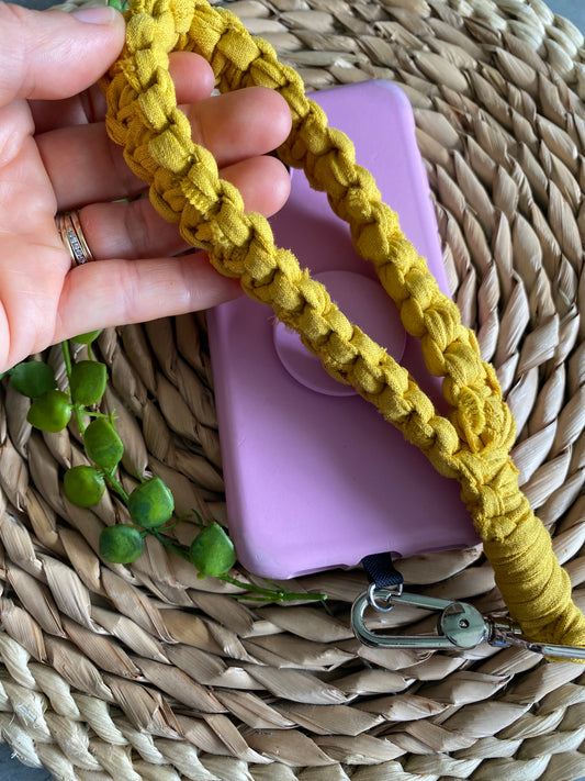 Handmade macrame phone wrist strap, made with mustard yellow T shirt yarn, and a metal clip. Attached to a pink phone case with a universal tether. 