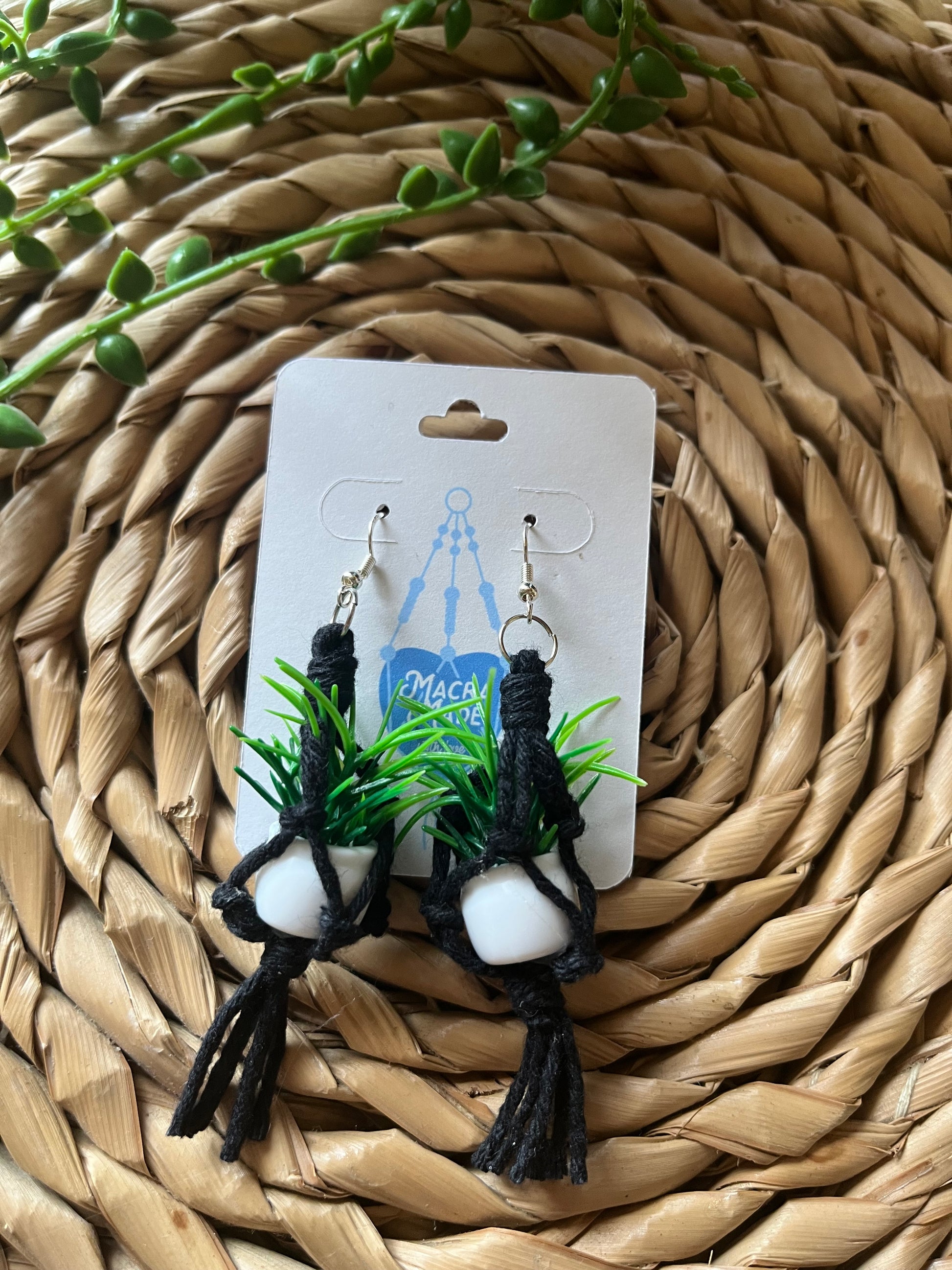 A pair of mini macrame plant hanger earrings made with black yarn and small artificial plants, with silver hooks, on a white backing card, on a wicker surface. 