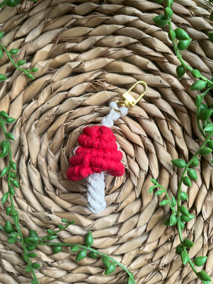 A handmade macrame mushroom keyrings, made with eco friendly recycled cotton in red, with a shiny metal clasp. The keyring is displayed on a wicker mat, with some long green plant vines.