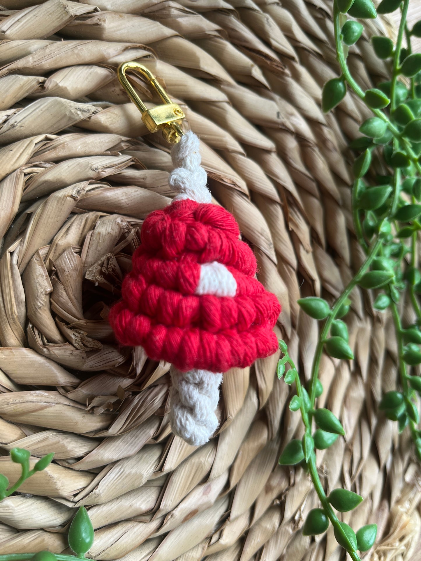 A handmade macrame mushroom keyring, made with eco friendly recycled cotton in red, with a shiny metal clasp. The keyring is displayed on a wicker mat, with some long green plant vines.