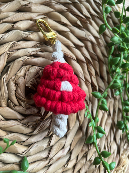 A handmade macrame mushroom keyring, made with eco friendly recycled cotton in red, with a shiny metal clasp. The keyring is displayed on a wicker mat, with some long green plant vines.