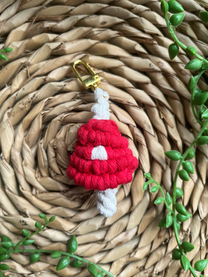 A handmade macrame mushroom keyring, made with eco friendly recycled cotton in red, with a shiny metal clasp. The keyring is displayed on a wicker mat, with some long green plant vines.