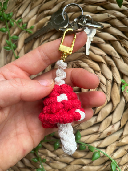 A hand is holding a handmade macrame mushroom keyring, made with eco friendly recycled cotton in red, with a shiny metal clasp. The keyring is displayed on a wicker mat, with some long green plant vines attached to a small set of silver keys.
