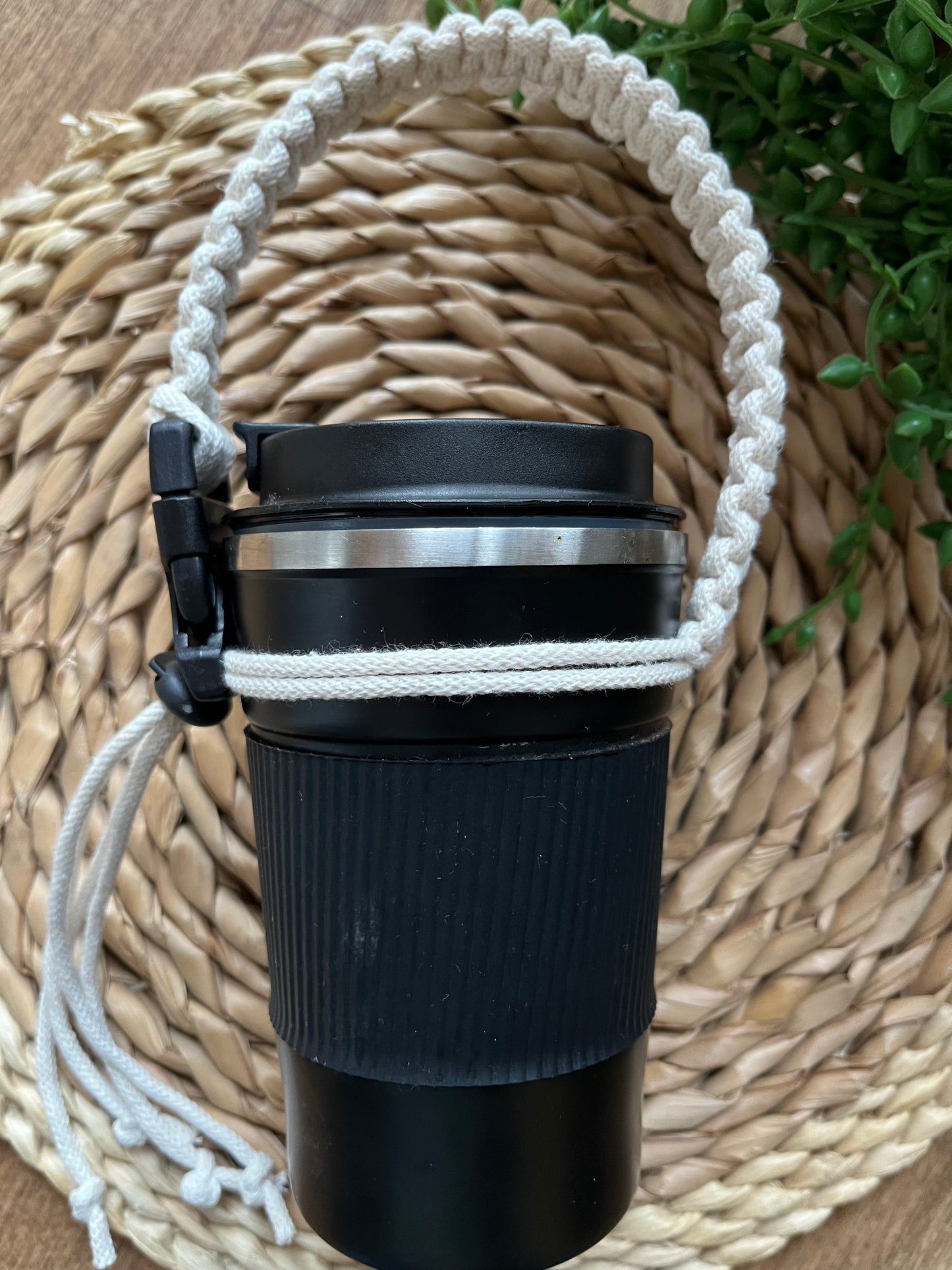 A black reusable coffee cup is displayed on a wicker mat, with some green plant vines, with a short natural adjustable macrame coffee cup holder strap attached to it. 