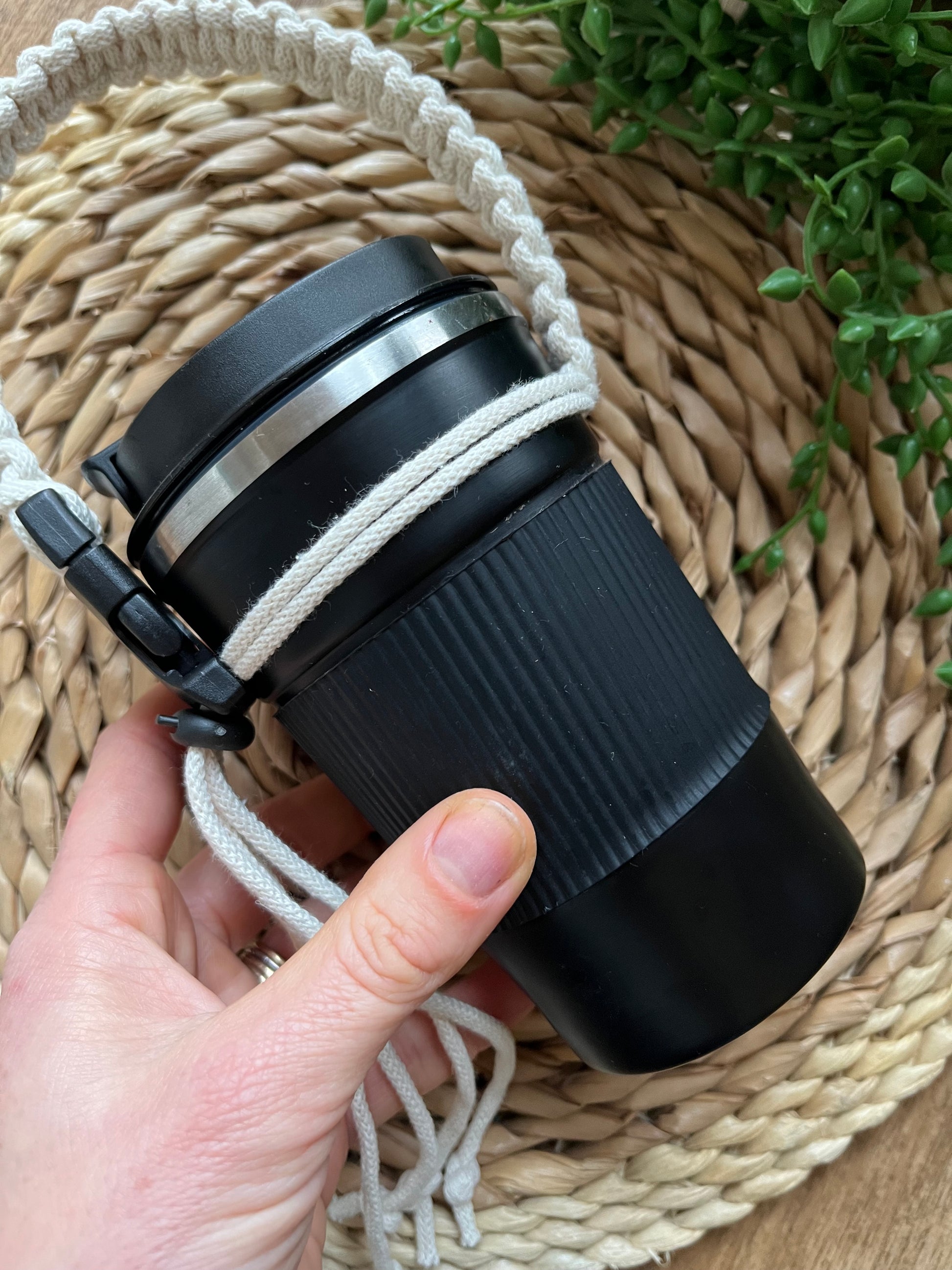 A hand holds a black reusable coffee cup, which has a handmade macrame cup holder strap attached to it, with a black buckle and toggle. 