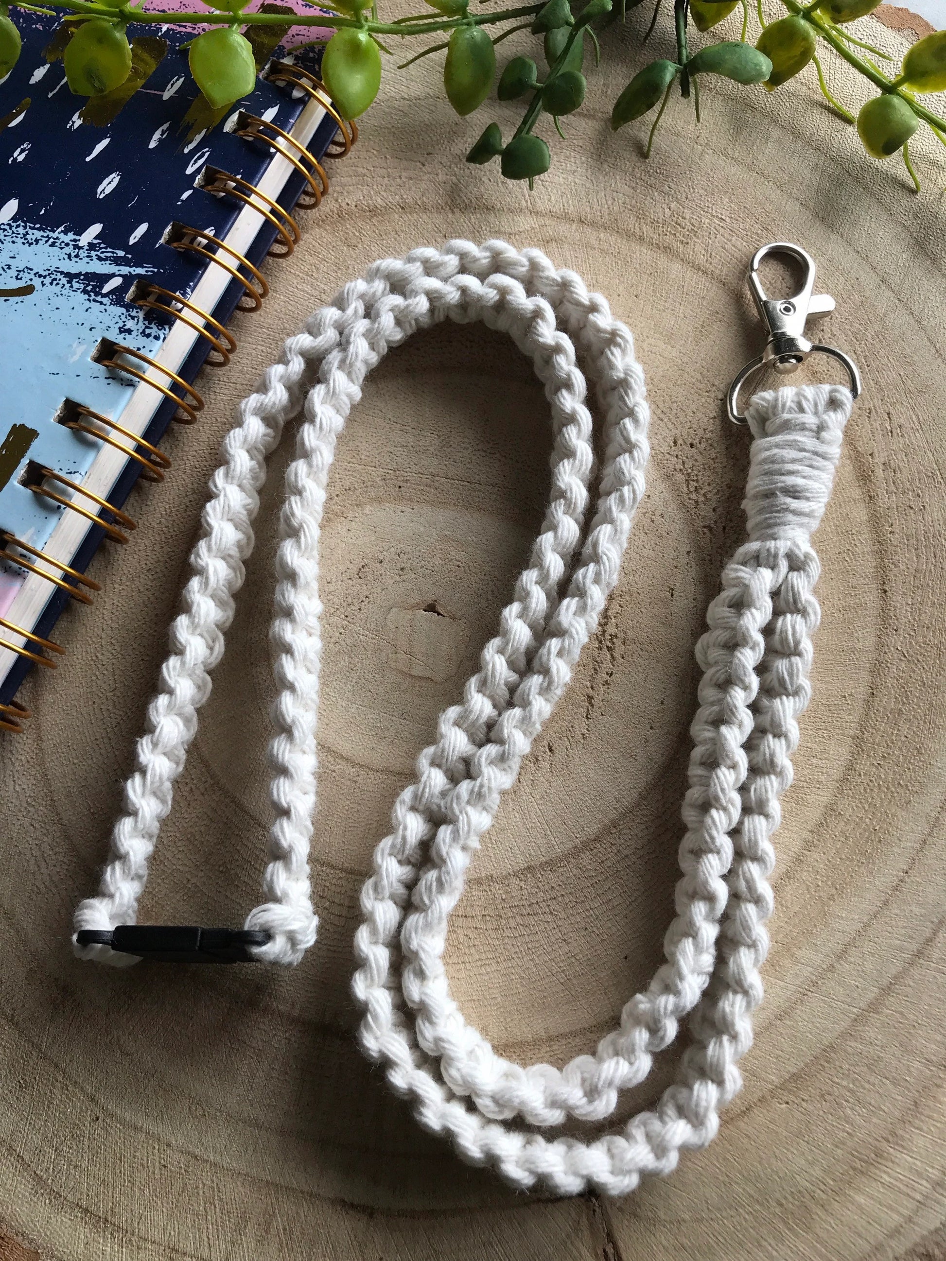 A handmade, eco-friendly lanyard lies on a rustic wooden surface. The white crocheted lanyard features a metal clasp at one end and a black plastic buckle at the other. Nearby is a spiral-bound notebook with a colorful cover and green foliage. This **Eco friendly lanyard badge holder - 34” by Macra-Made-With-Love** is perfect for carrying your essentials while staying environmentally conscious.