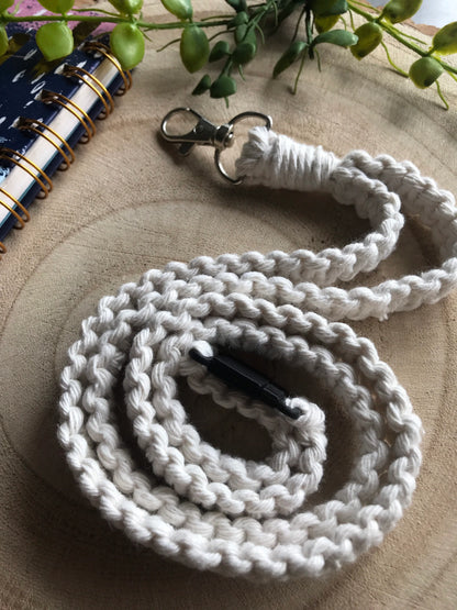 A handmade, braided white cord lanyard with a lobster clasp on one end and a black closure on the other, coiled neatly on a wooden surface next to a spiral-bound notebook with a black cover. Green plant leaves are visible in the background. Perfect as an eco-friendly ID badge holder, introducing the Eco friendly lanyard badge holder - 34” by Macra-Made-With-Love.