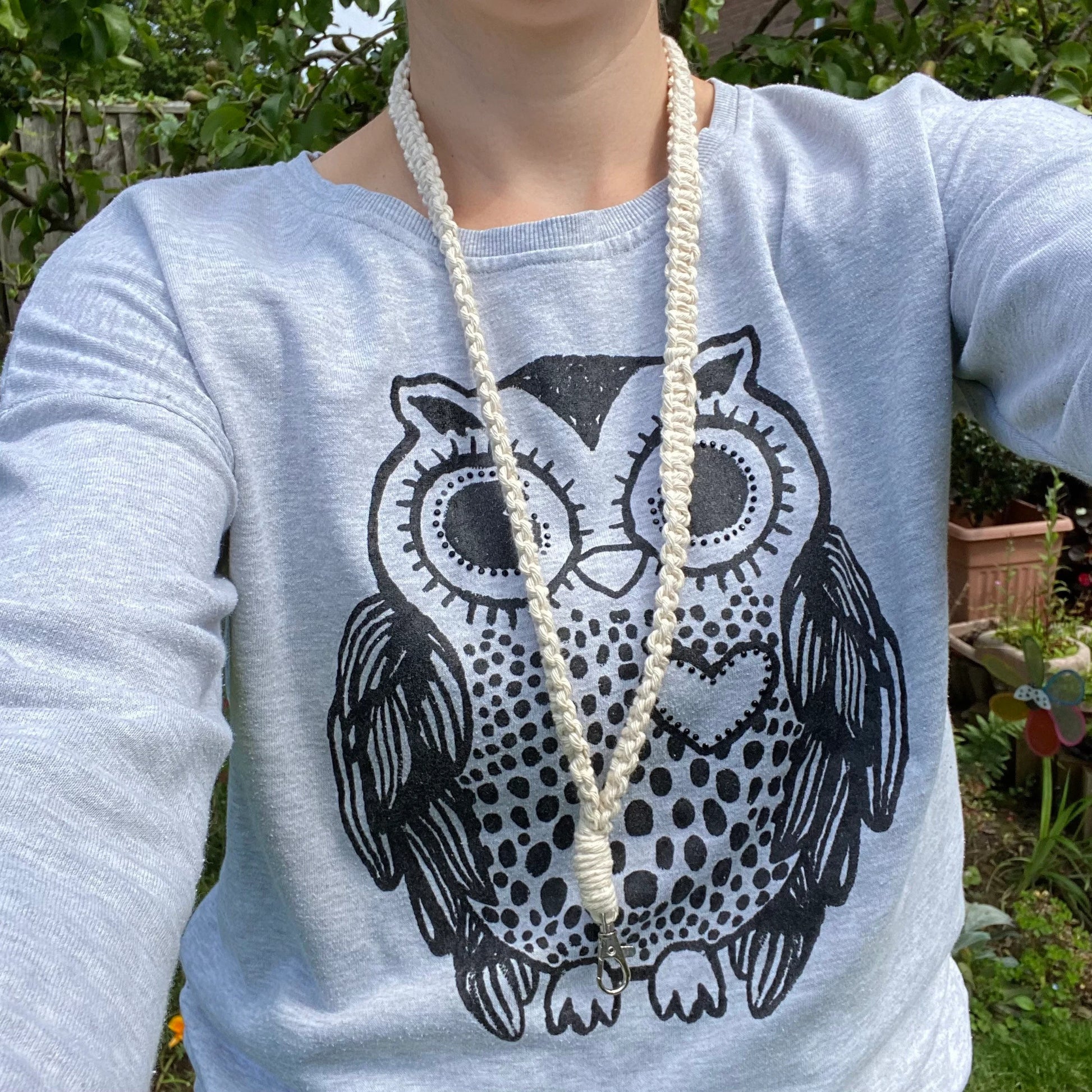 A person wearing a light grey sweatshirt with a black graphic of an owl on it and a long, crocheted white necklace that doubles as an **Eco friendly lanyard badge holder - 34”** by **Macra-Made-With-Love**. The background features greenery and a garden with some flowers, suggesting the photo was taken outdoors on a sunny day.