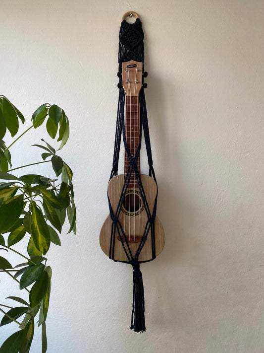 A light brown ukulele sits inside of a handmade macrame wall hanging ukulele hanger, which is made with navy blue recycled cotton, and a repurposed wooden hanging ring. A large green leafy plant is to the left of the image. 