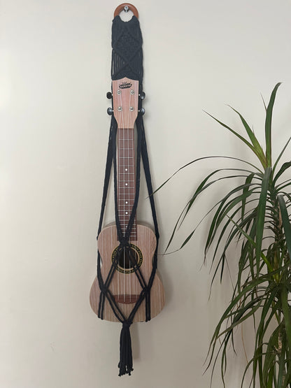 A light brown ukulele sits inside of a handmade macrame wall hanging ukulele hanger, which is made with navy blue recycled cotton, and a repurposed wooden hanging ring. A large green leafy plant is to the right of the image. 