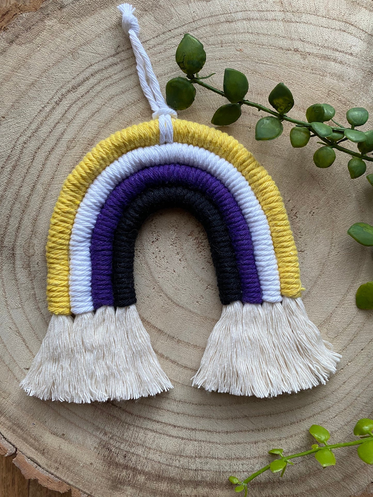 A small macrame rainbow made with yellow white purple and black yarns to represent the colours of the non binary pride flag, with a white hanging loop, placed on a wooden surface with green plant vines. 