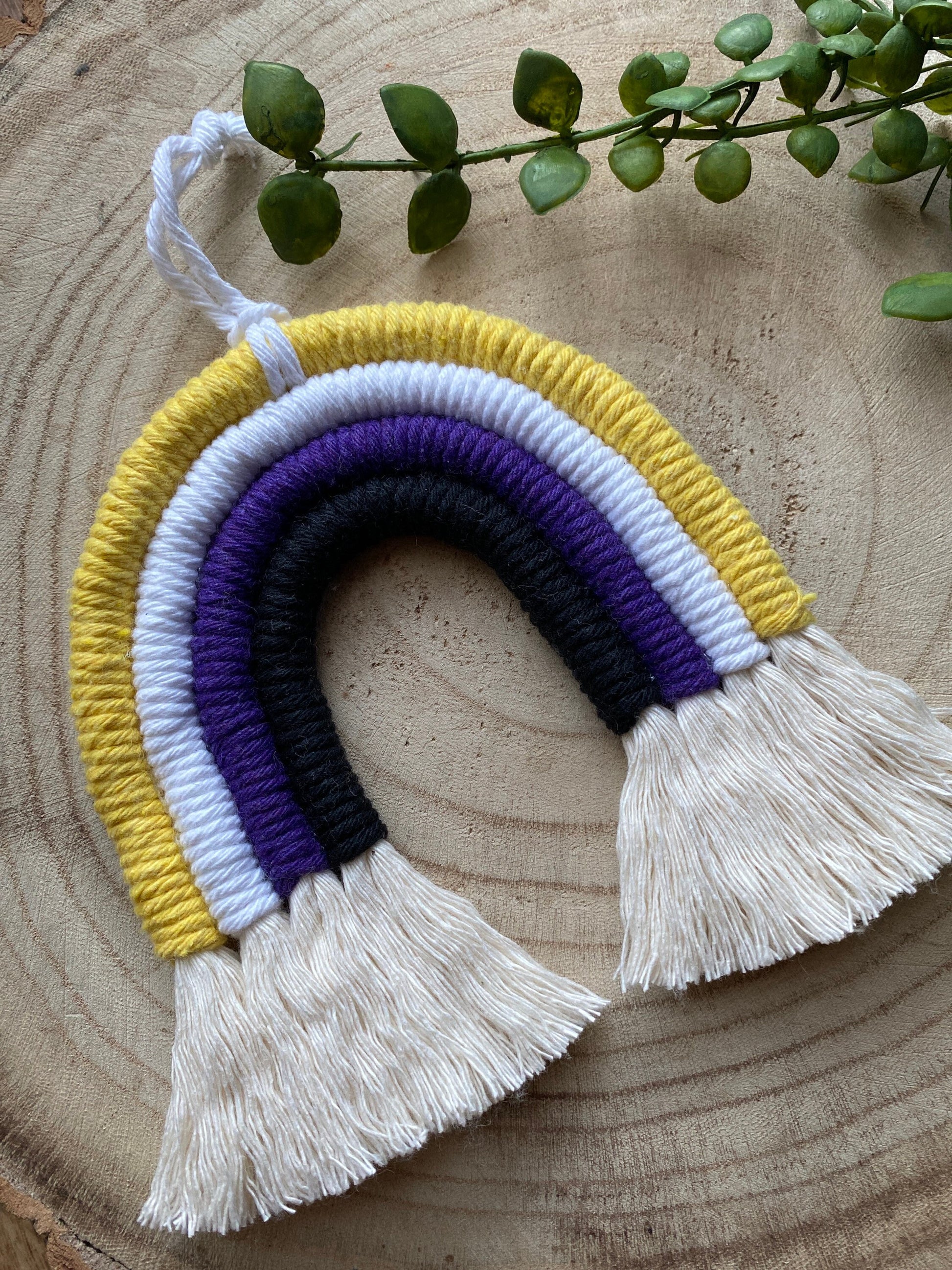 A small macrame rainbow made with yellow white purple and black yarns with white tassels to represent the colours of the non binary pride flag, with a white hanging loop, placed on a wooden surface with green plant vines. 