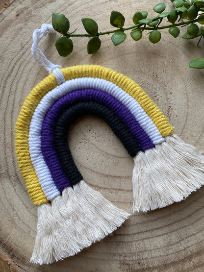 A small macrame rainbow made with yellow white purple and black yarns with white tassels to represent the colours of the non binary pride flag, with a white hanging loop, placed on a wooden surface with green plant vines. 