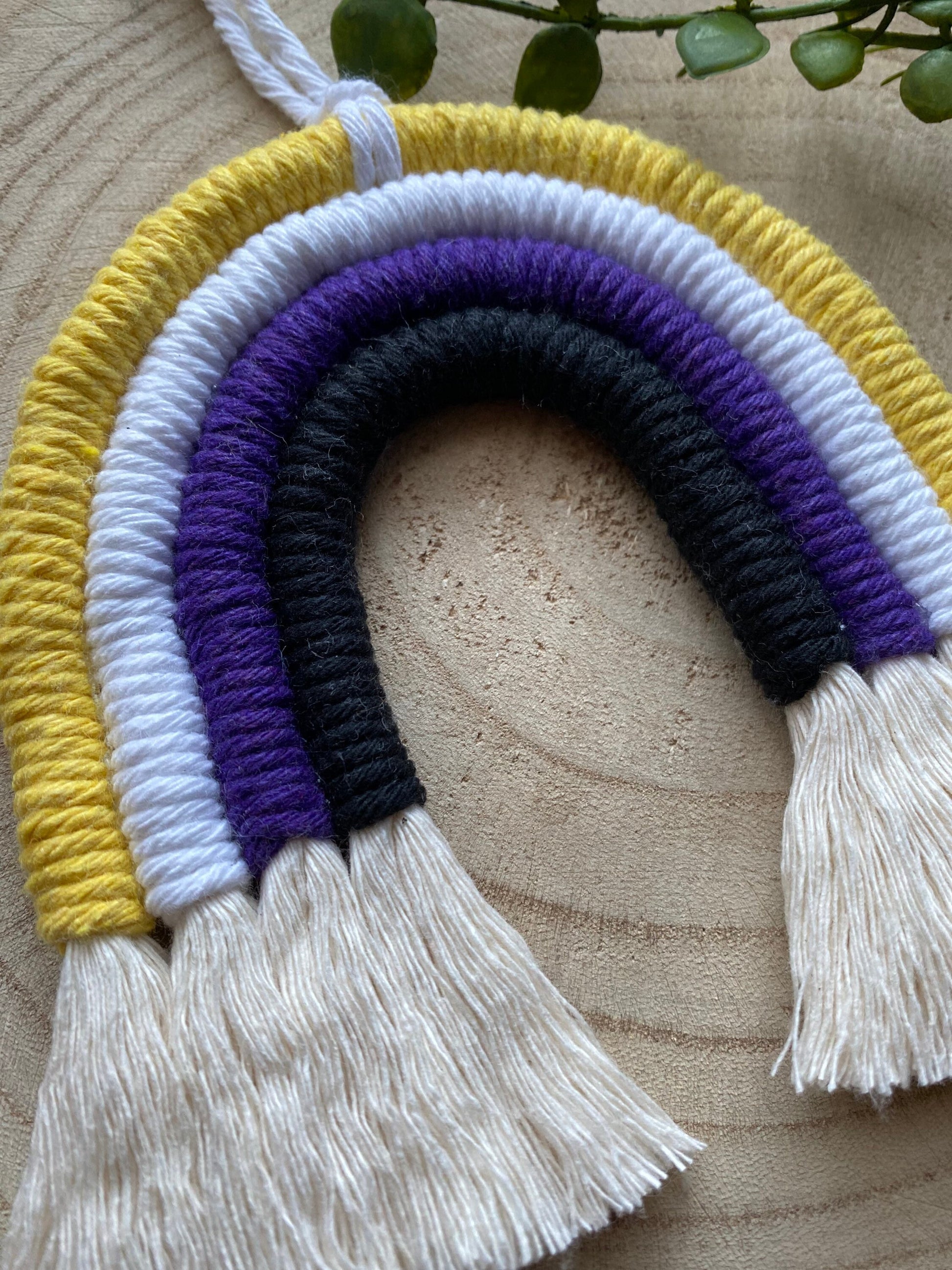 Close up of a small macrame rainbow made with yellow white purple and black yarns to represent the colours of the non binary pride flag, with long white tassels at the bottom. 