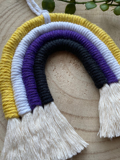 Close up of a small macrame rainbow made with yellow white purple and black yarns to represent the colours of the non binary pride flag, with long white tassels at the bottom. 