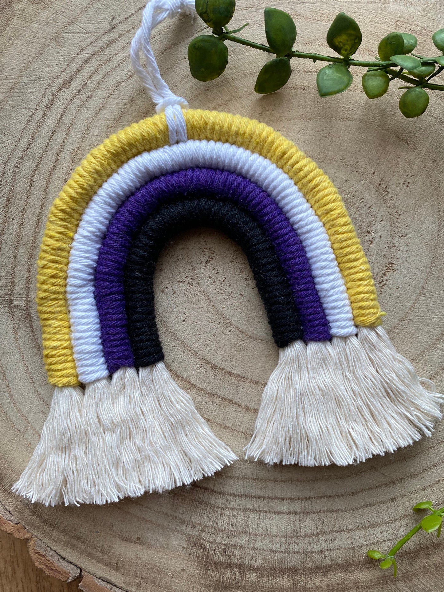 A small macrame rainbow made with yellow white purple and black yarns to represent the colours of the non binary pride flag, with a white hanging loop, placed on a wooden surface with green plant vines. 