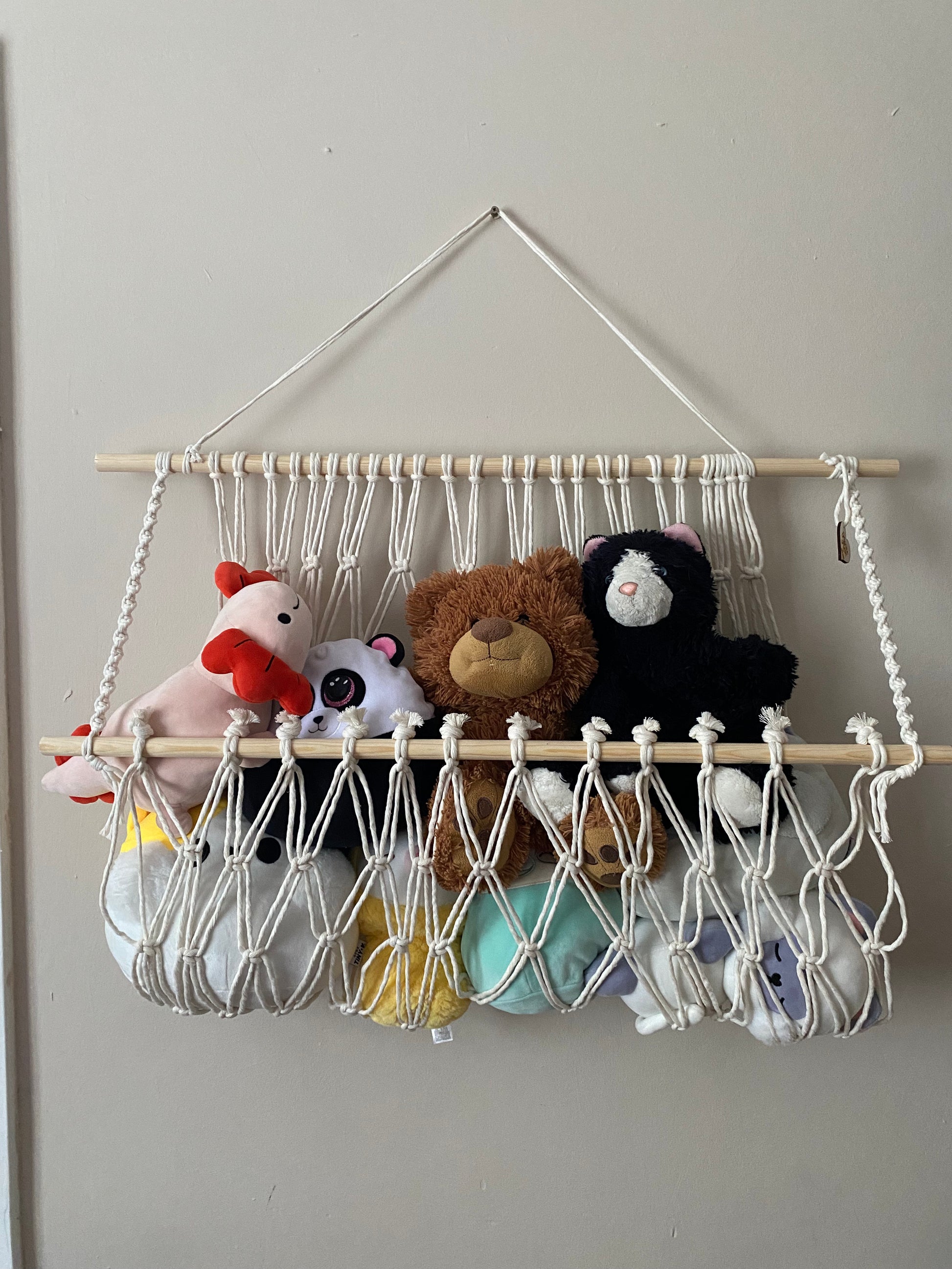 A macrame toy hammock made with natural yarn and wooden dowels, hanging against a beige wall, filled with soft toys and teddies. 