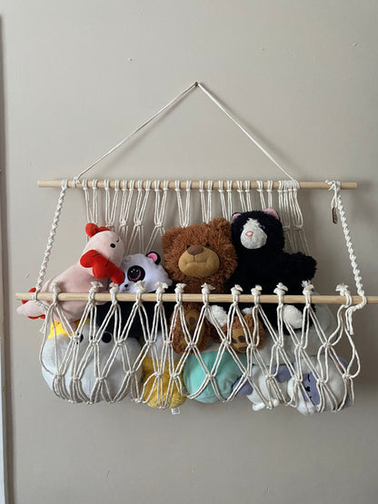 A macrame toy hammock made with natural yarn and wooden dowels, hanging against a beige wall, filled with soft toys and teddies. 