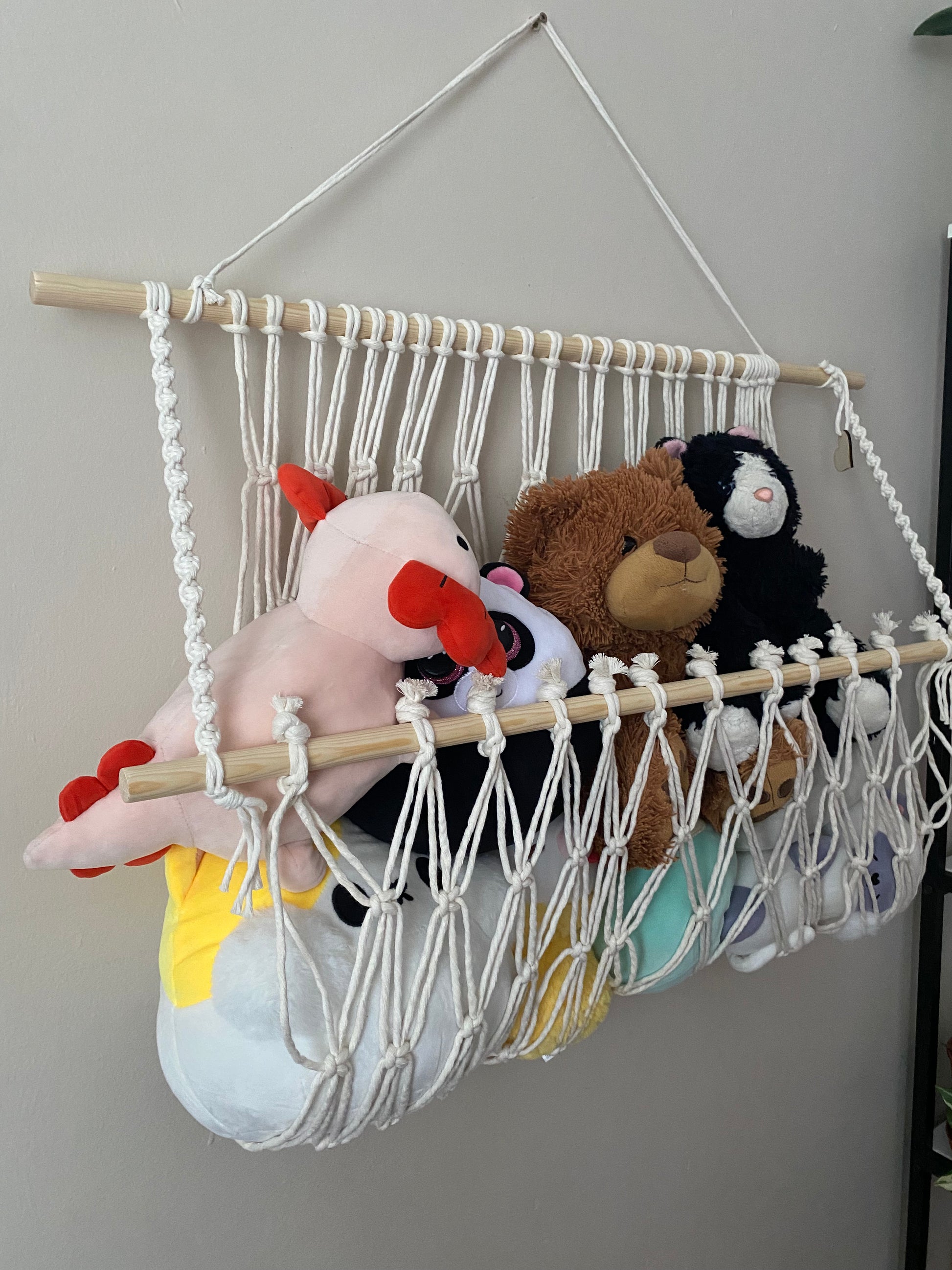 A macrame toy hammock made with natural yarn and wooden dowels, hanging against a beige wall, filled with soft toys and teddies. 