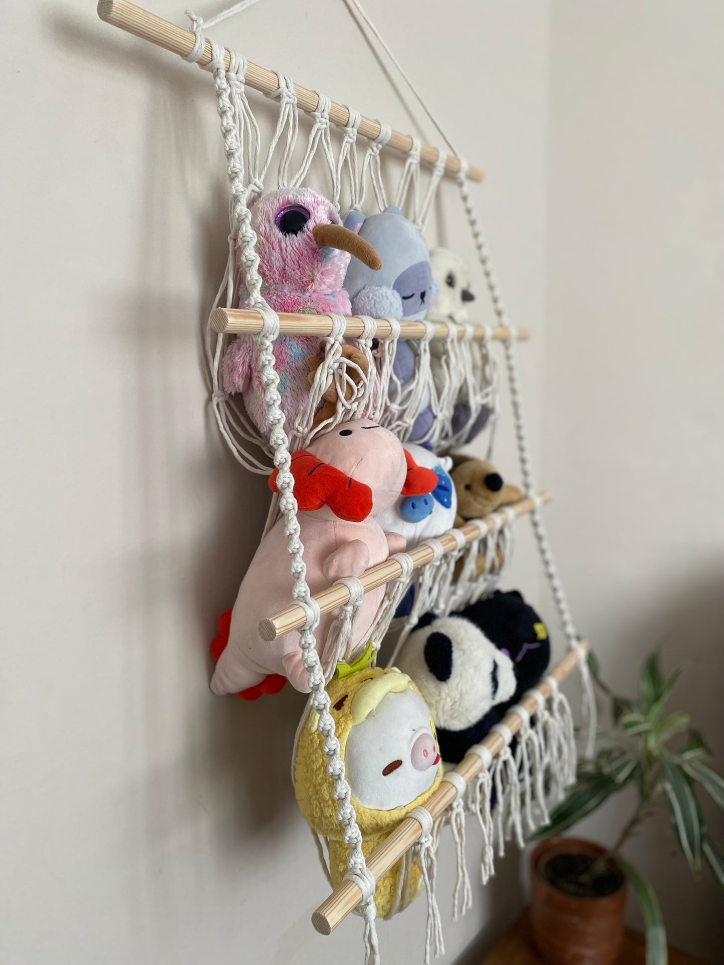 A close up side view of a large macrame toy hammock made with natural yarn and four wooden dowels, hanging against a beige wall, filled with soft toys and teddies. 