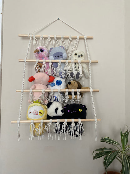 A macrame toy hammock with three layers, made with natural yarn and wooden dowels, hanging against a beige wall, filled with soft toys and teddies. 